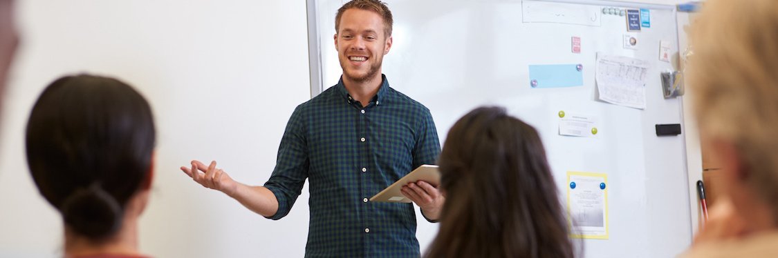 Junger Lehrer steht vor seiner Klasse, im Hintergrund ist ein Whiteboard zu sehen.